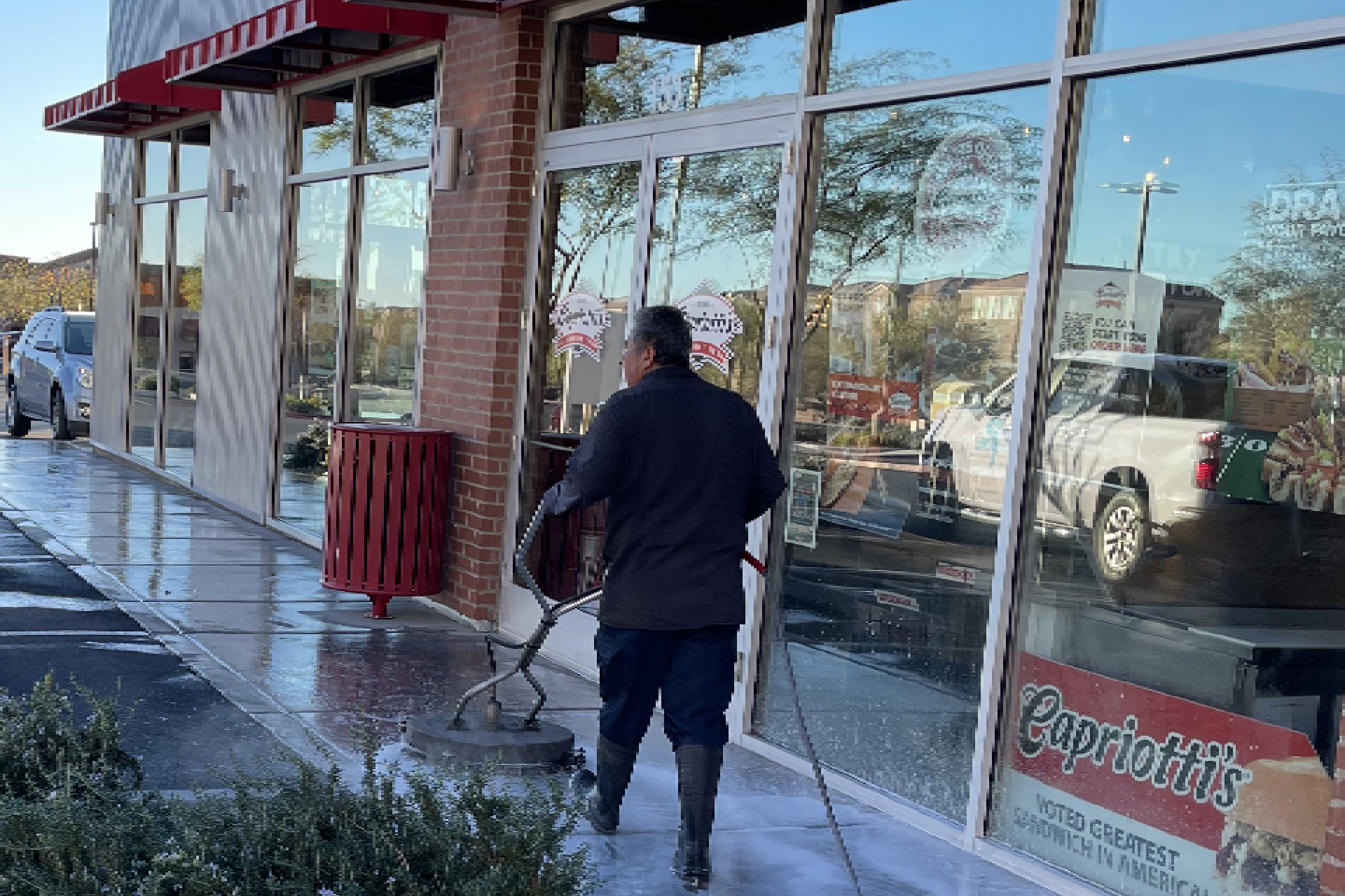 a man wearing a black jacket and boots, standing outside a building and using a pressure washer to clean the sidewalk